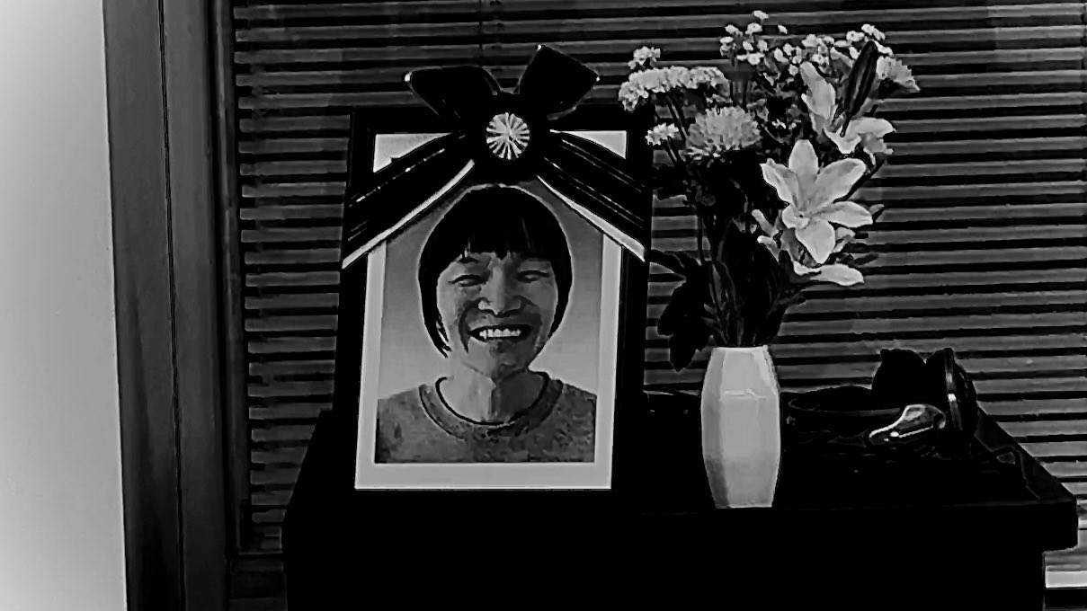 写真の女性の遺影と花瓶の花、黒い靴が置かれた仏壇が写っています。