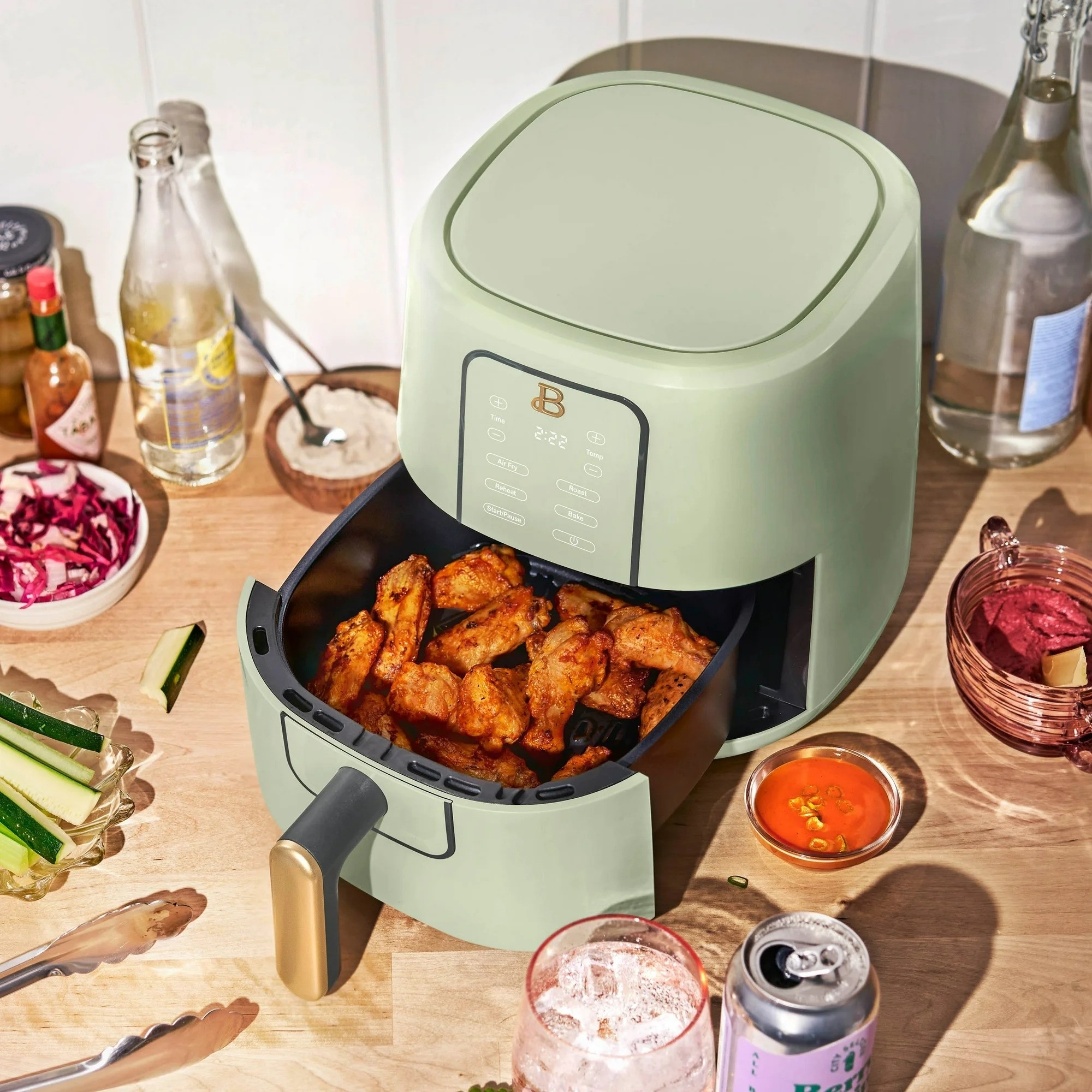 A green air fryer with its tray pulled out, displaying cooked chicken wings, on a kitchen counter among various condiments