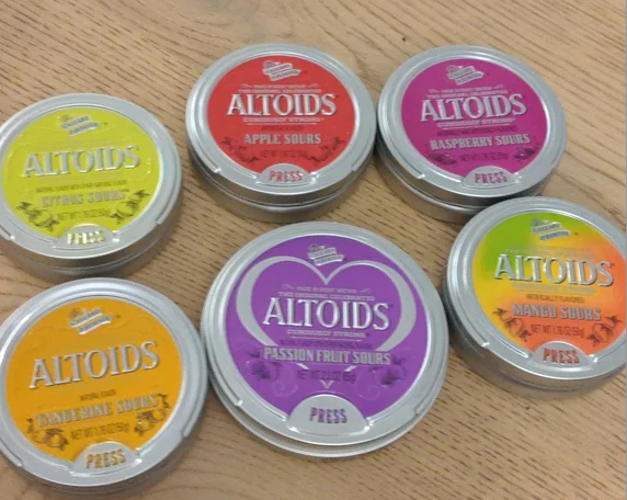 Six different flavors of Altoids Sours in circular tins arranged on a wooden surface