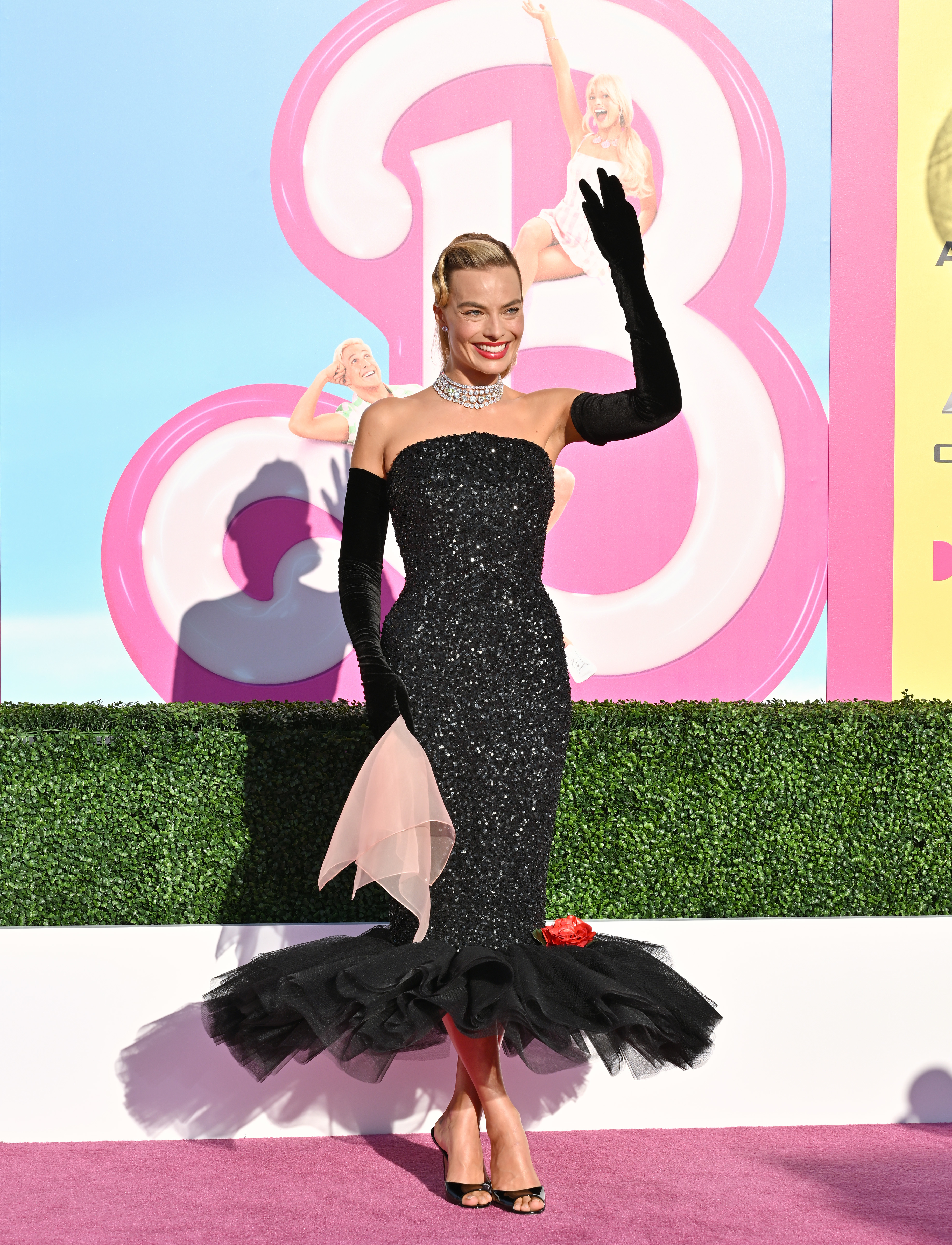 Margot Robbie in a black gown with ruffled detail, waving on the &#x27;Barbie&#x27; premiere backdrop