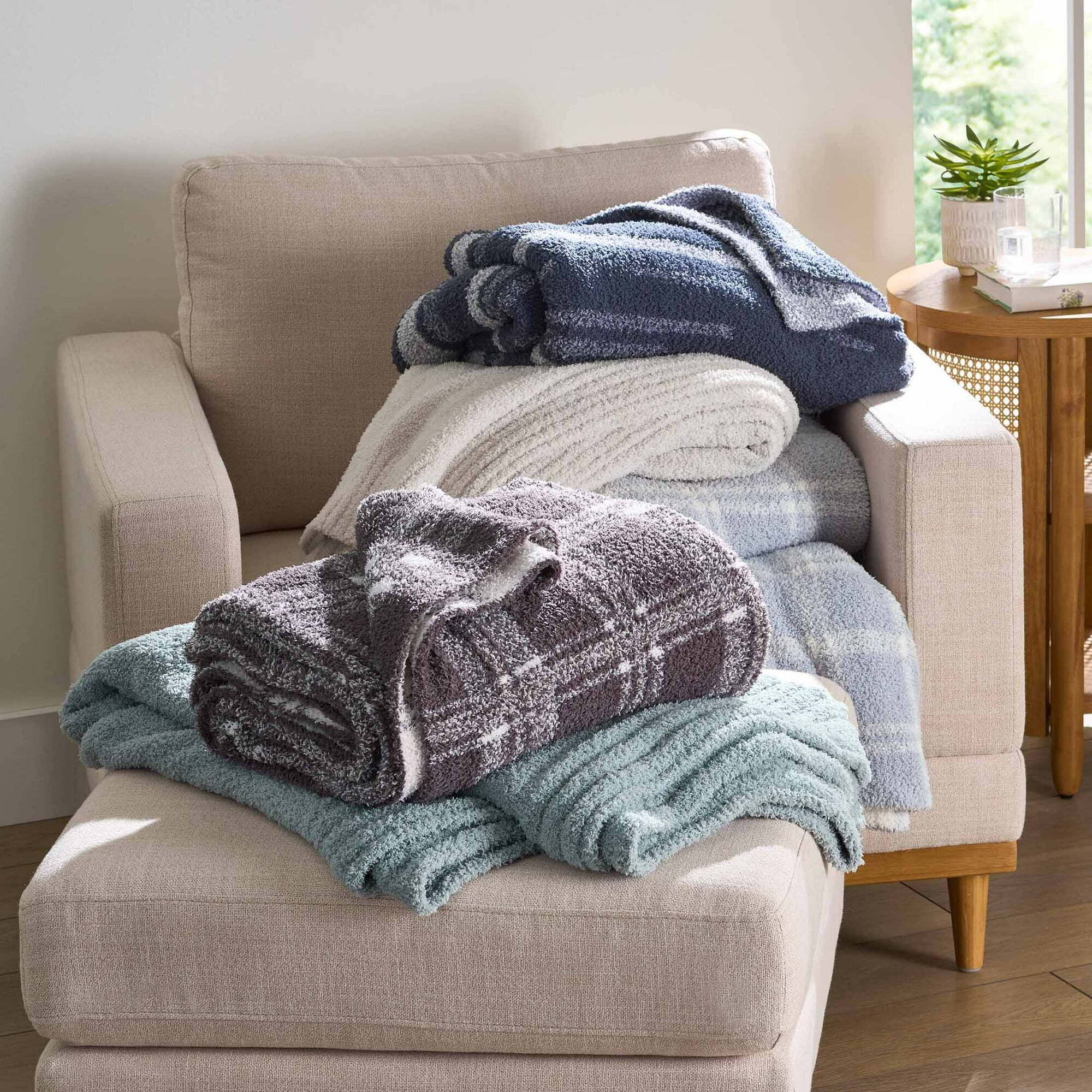 A stack of plush blankets in varied patterns on a beige armchair