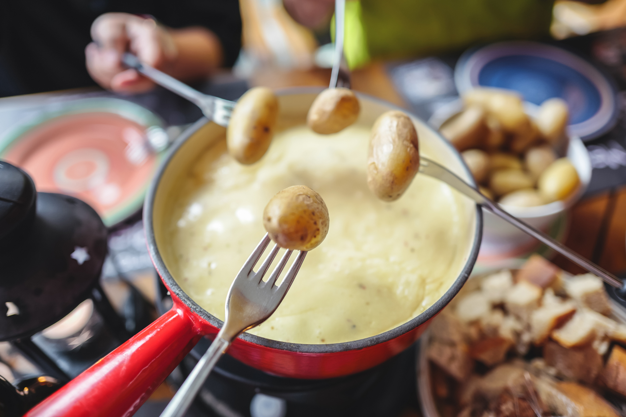 A person dipping a potato onto a fork into a pot of fondue with plates of food surrounding it