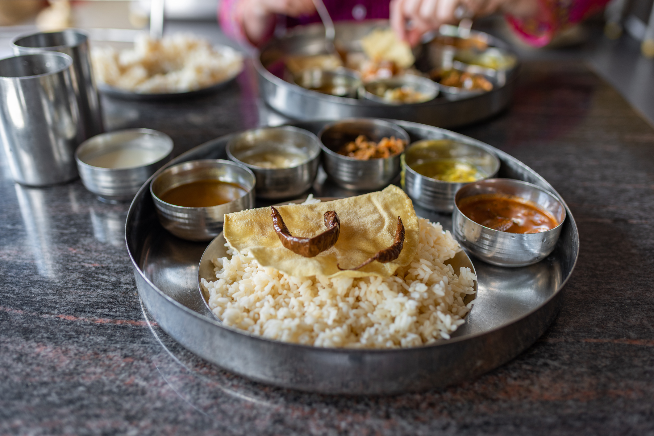 Indian thali with rice, various curries, and a papadum on a metal tray, hands serving food in background