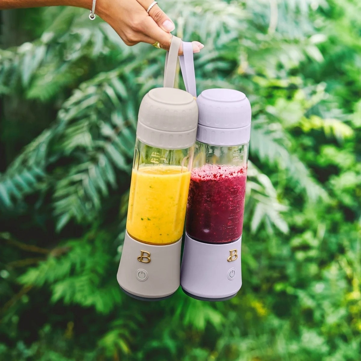 Hand holding two portable blenders with yellow and red smoothies against a backdrop of green leaves
