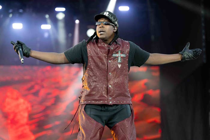 Music artist in a maroon vest and black cap performs on stage with arms outstretched