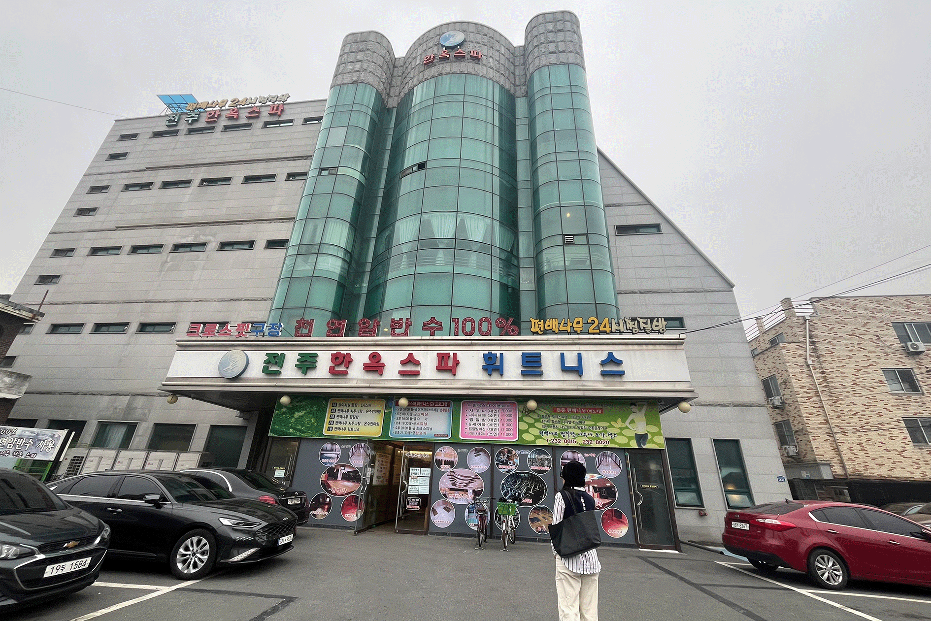 Exterior view of a building with signs, a person with backpack standing in front, parked cars visible