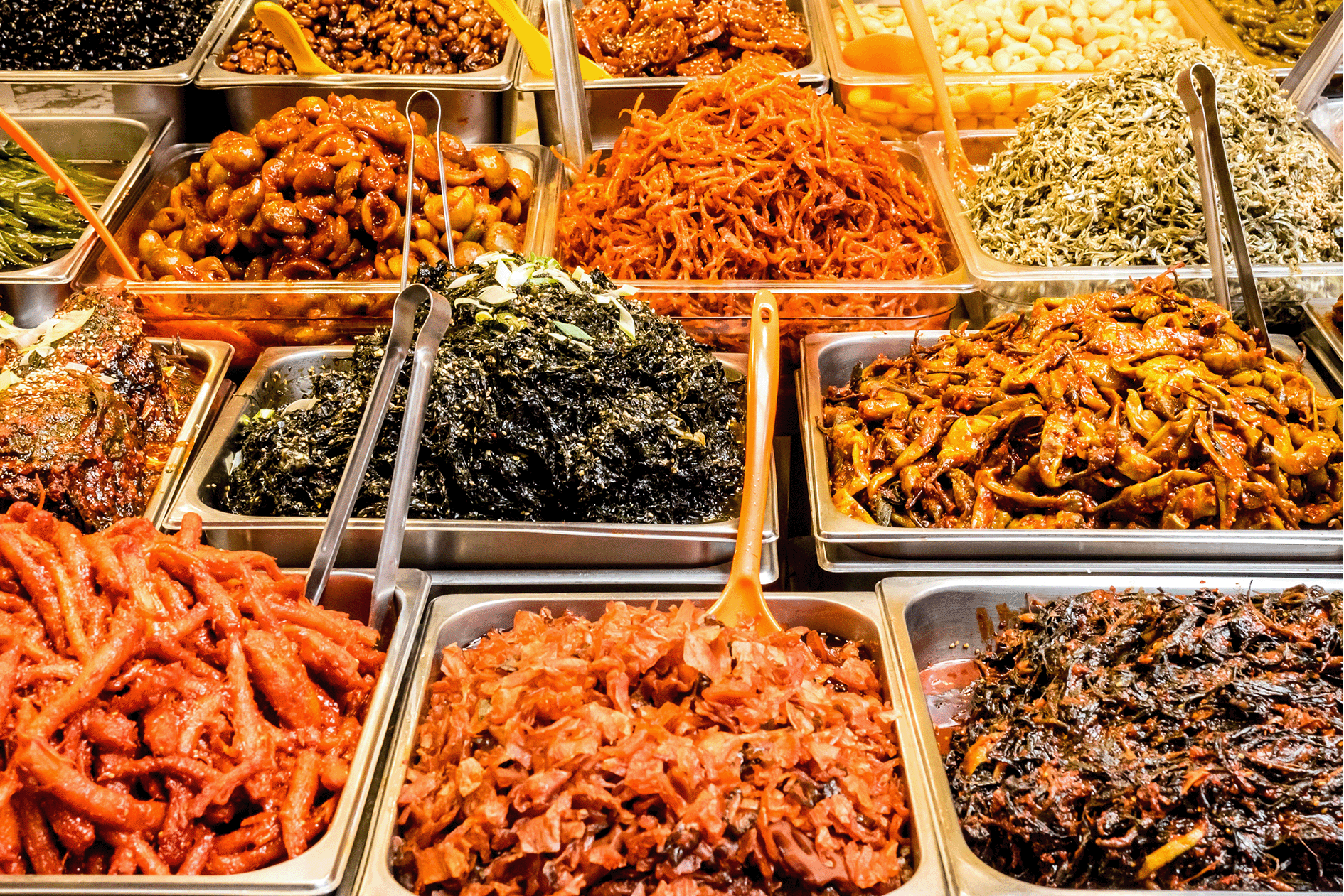Variety of Korean side dishes displayed in metal trays at a market