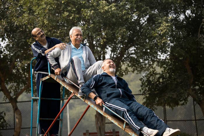 Three elderly men share a light moment on a seesaw at a park, smiling and enjoying each other&#x27;s company