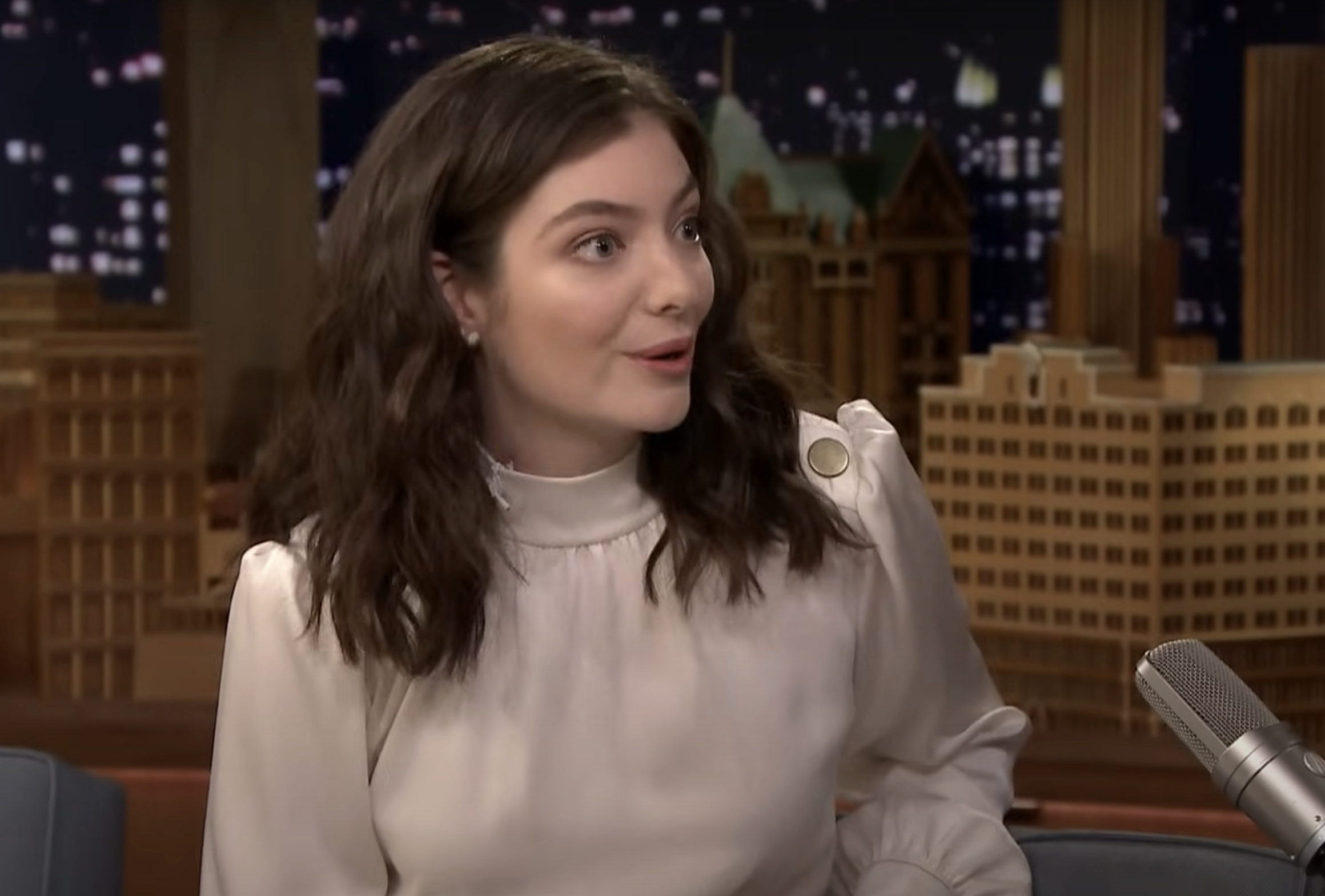 Lorde in light top on talk show, city backdrop, engaged in conversation