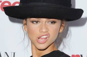 Person wearing a black hat and light makeup, biting their lower lip, at an event with logo backdrop