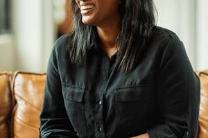 Woman in a black shirt and jeans sits smiling on a brown leather couch