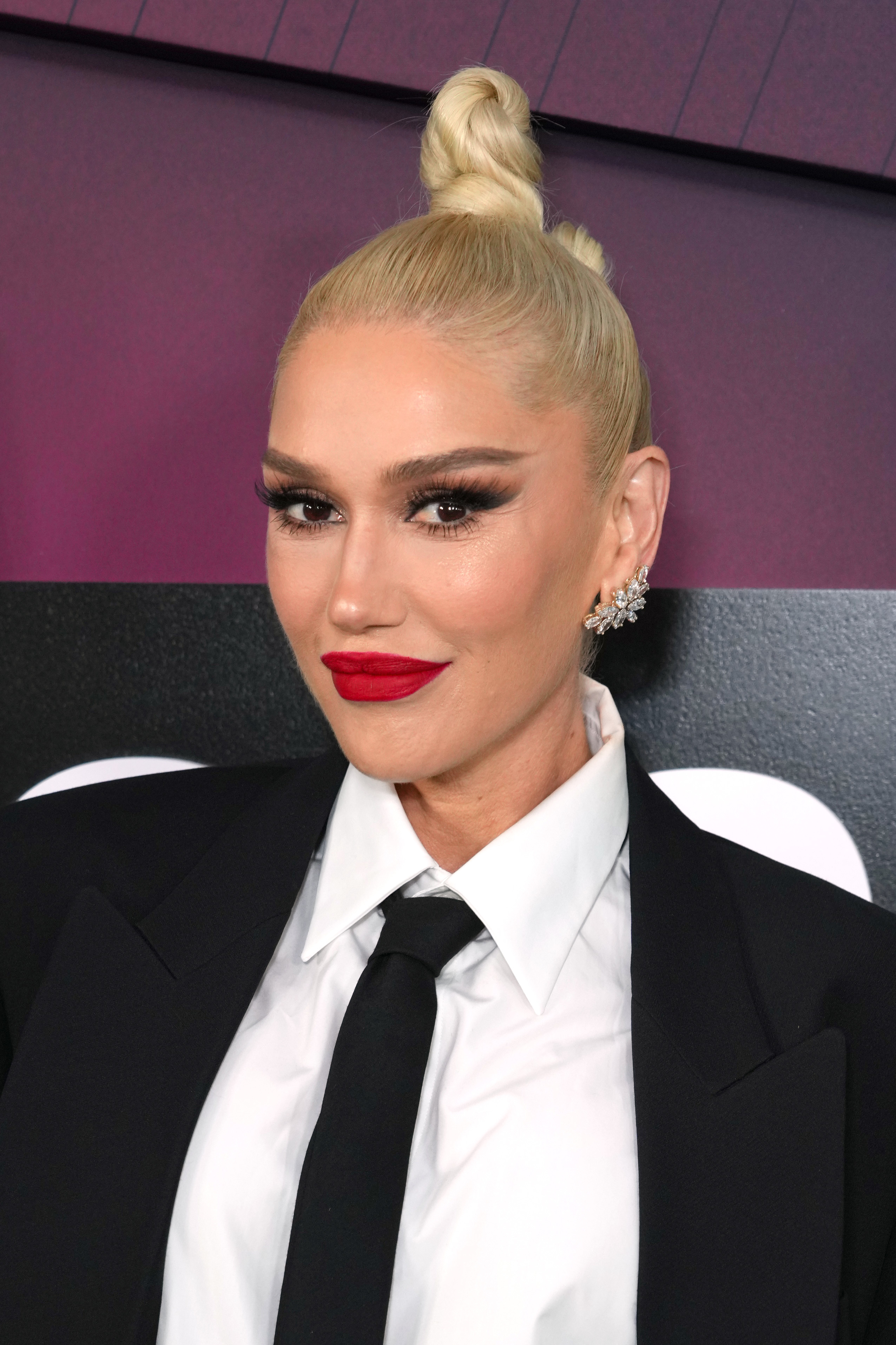 Gwen Stefani wearing a suit with a white shirt, black tie, and top bun hairstyle