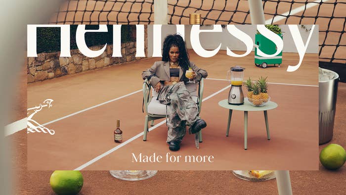 Woman lounging on a tennis court, with a drink, surrounded by Hennessy branding and snacks, in a stylish outfit