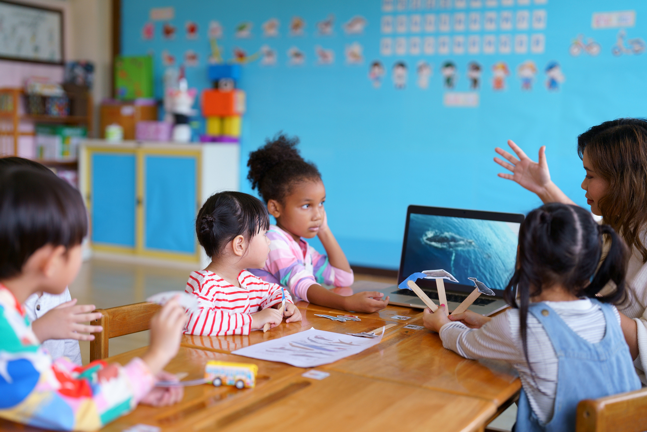 Teacher interacting with young students at a classroom table with a laptop and educational materials