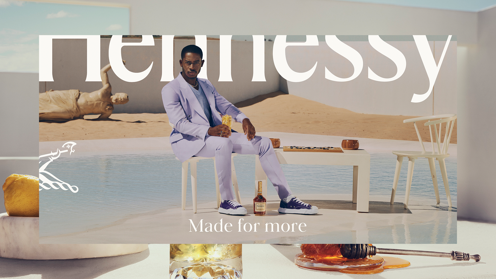 Man in a suit sitting by a pool with a glass and bottle of Hennessy, desert backdrop, ad text overlay