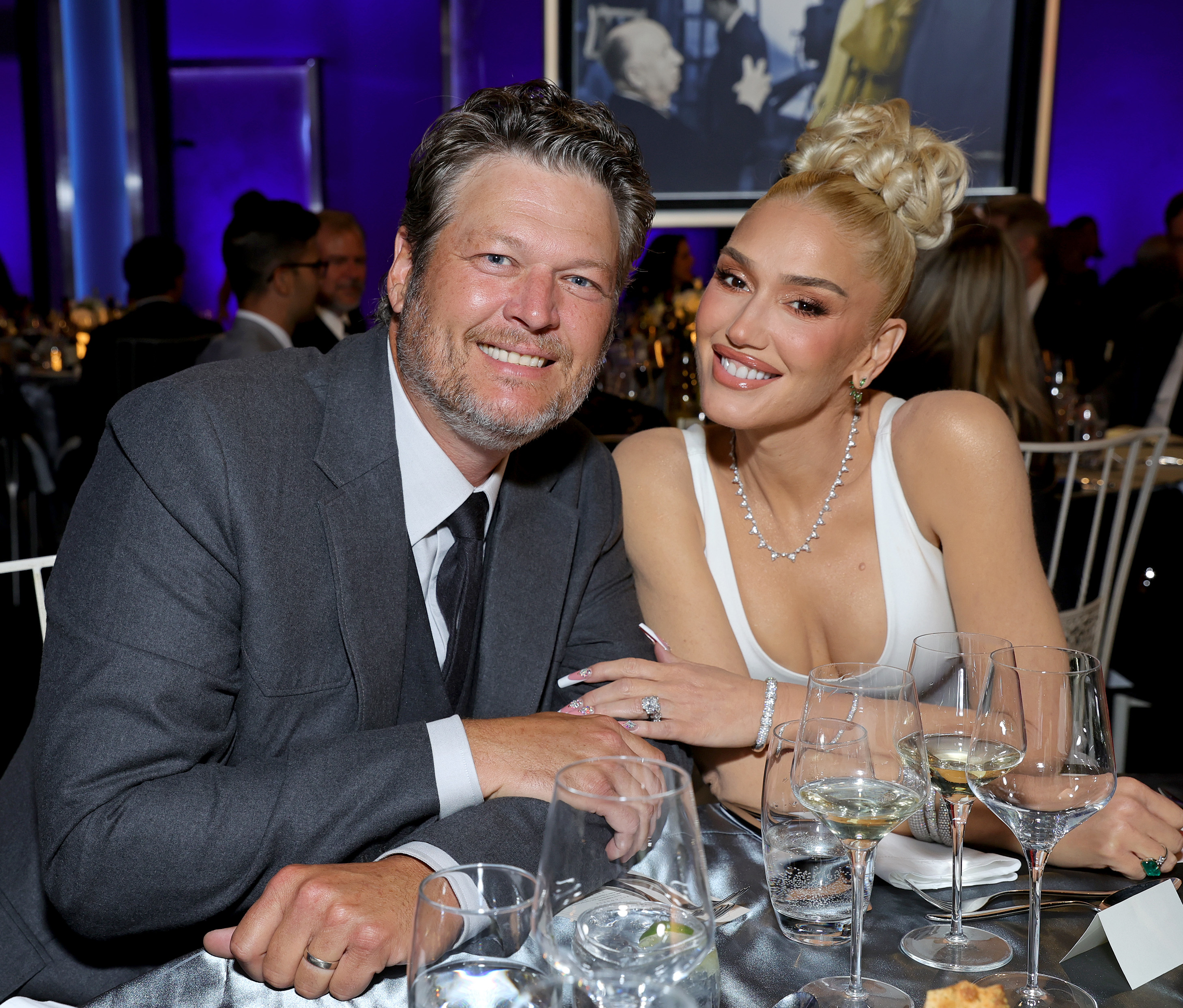 Blake Shelton and Gwen Stefani sitting together at a table with glasses in front. Gwen wears a white top and pearls, Blake in a suit