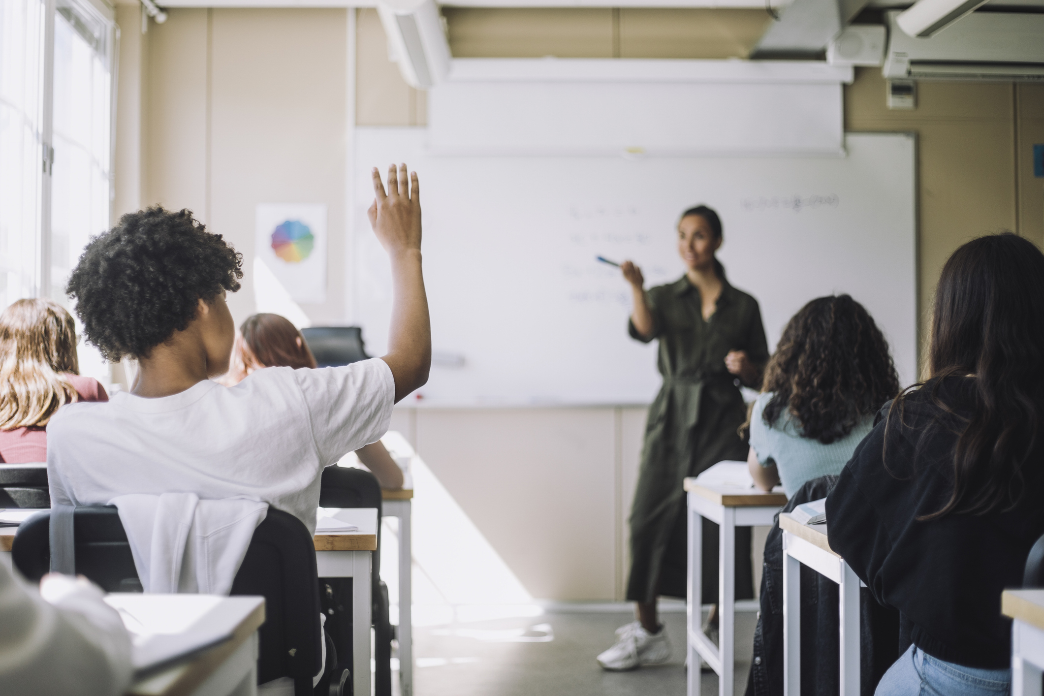 A student raises their hand in a classroom while the teacher engages with the class