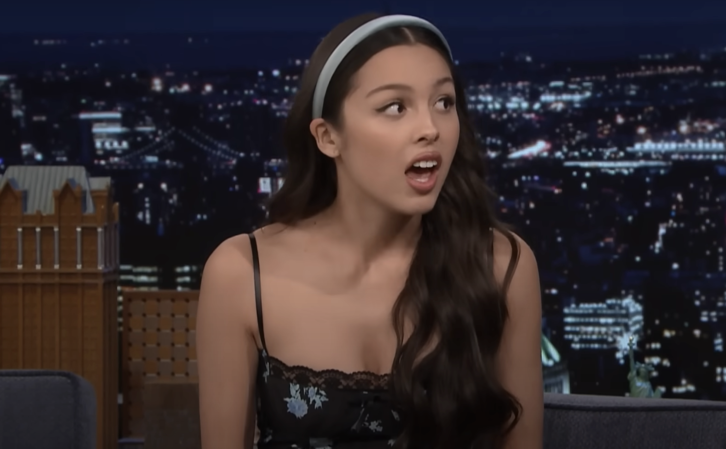Olivia wearing a headband and a lace-trimmed top sits on a talk show set, looking surprised mid-conversation