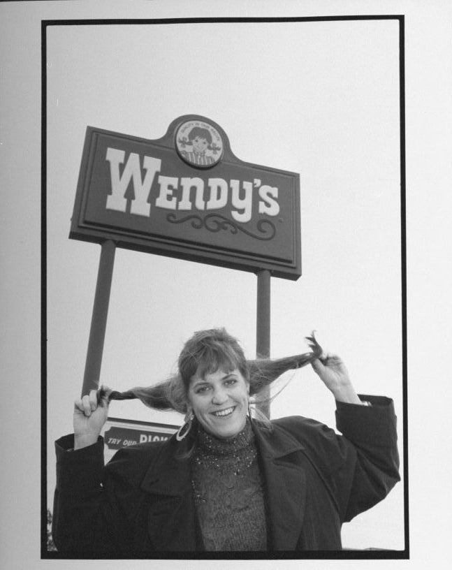 Woman smiling, holding her pigtails under Wendy&#x27;s sign, mimicking the logo&#x27;s character. She wears a jacket with a turtleneck