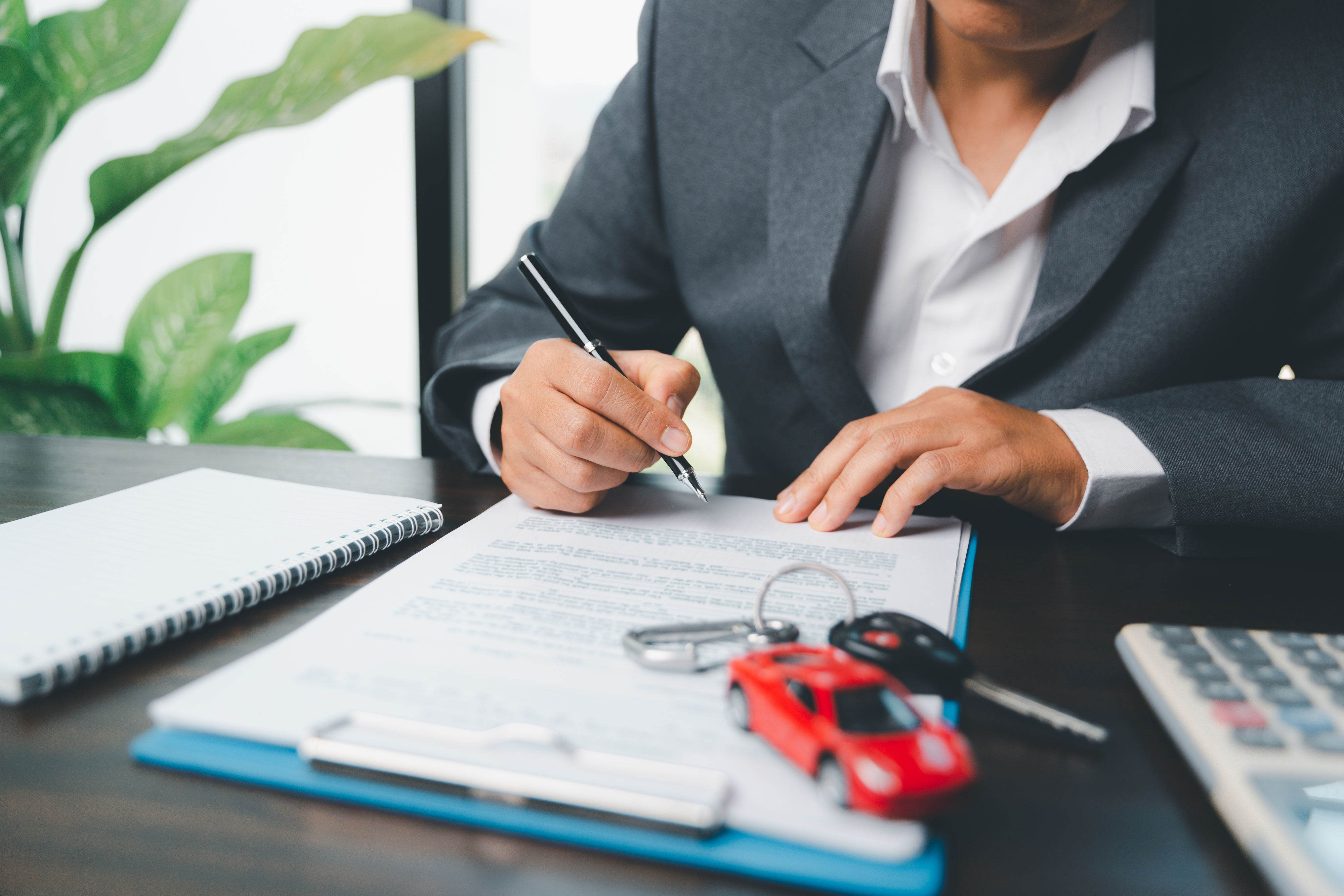 Person signing a document next to a small red car model, eyeglasses, and calculator on a desk