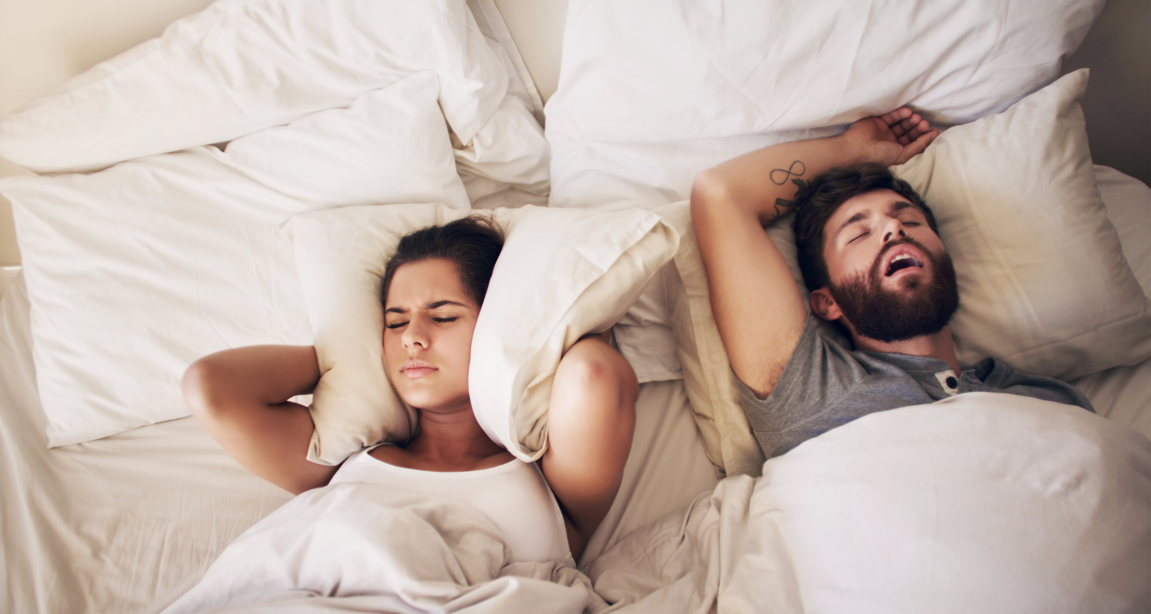 Two people lying in bed with one snoring and the other appearing frustrated
