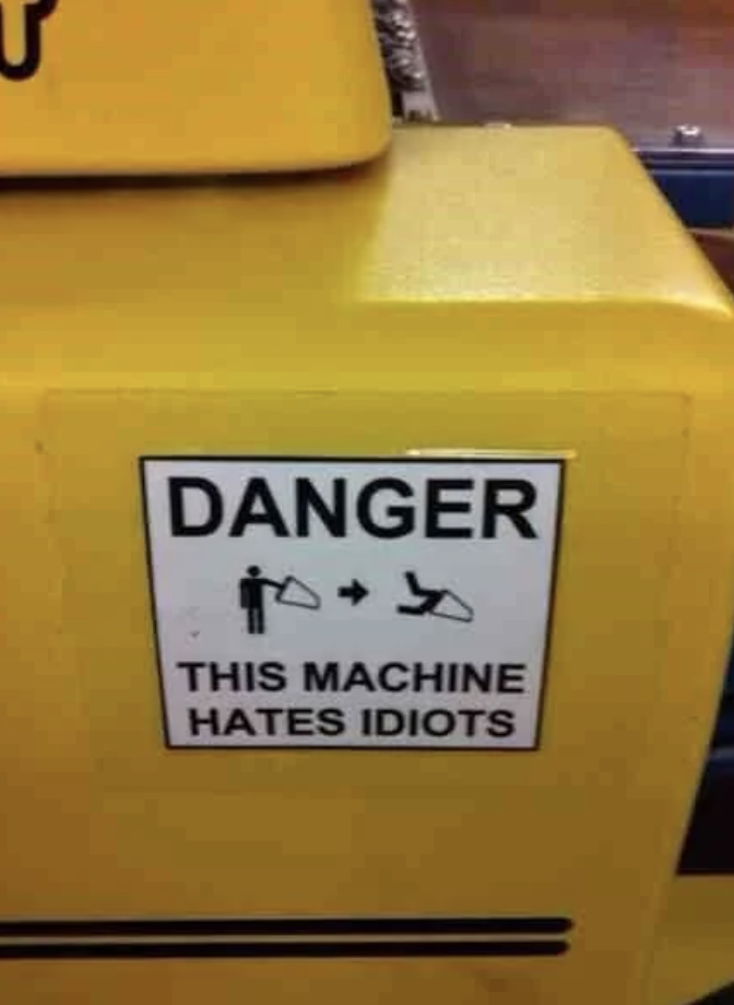 Warning sign on machinery humorously reading &quot;DANGER - THIS MACHINE HATES IDIOTS,&quot; with pictograms illustrating harm