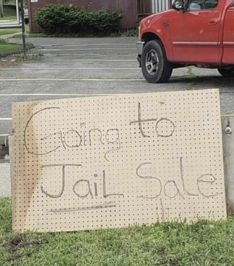 Handwritten sign reading &quot;Going to Jail Sale&quot; placed on grass near a sidewalk and vehicle