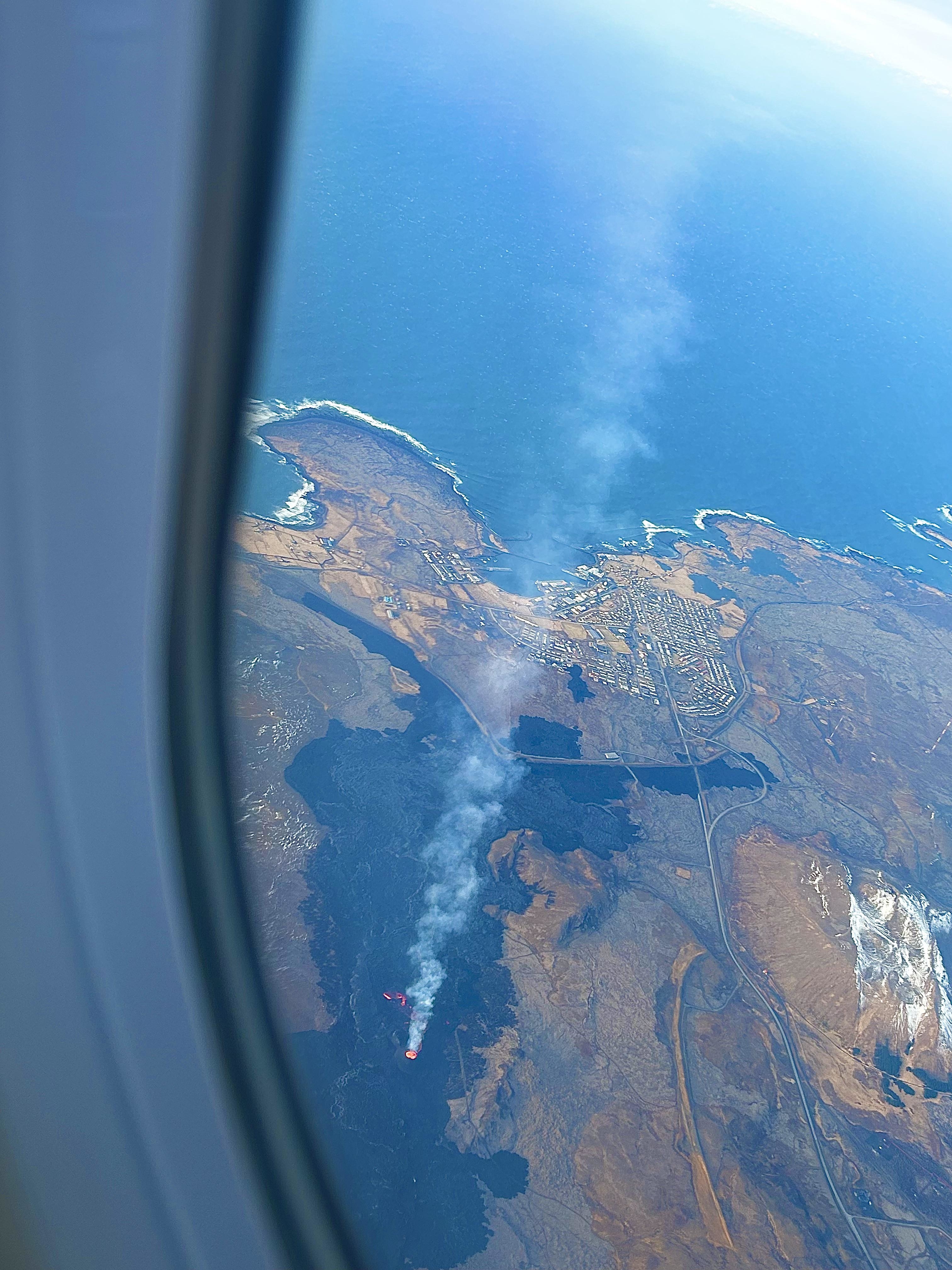 Aerial view of a coastline and a smoke plume from a fire, seen through an airplane window