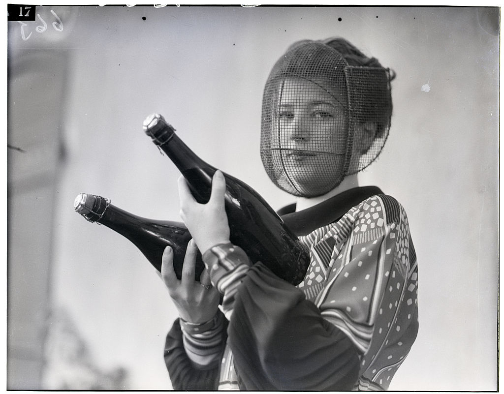Person in patterned attire holding champagne bottles, wearing a face net. Vintage setting