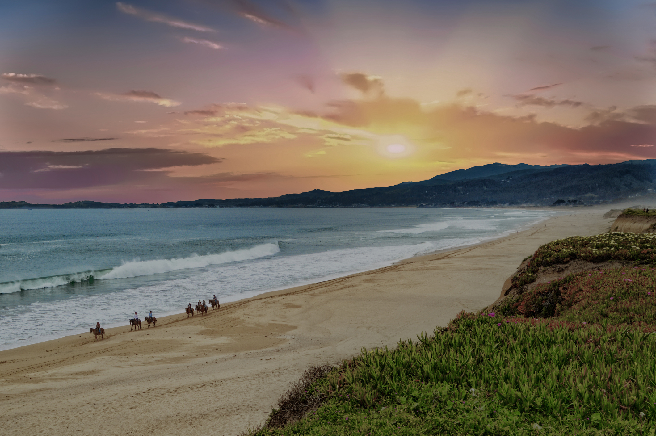 Sunset at a beach with people and horses walking along the shore, mountains in the distance