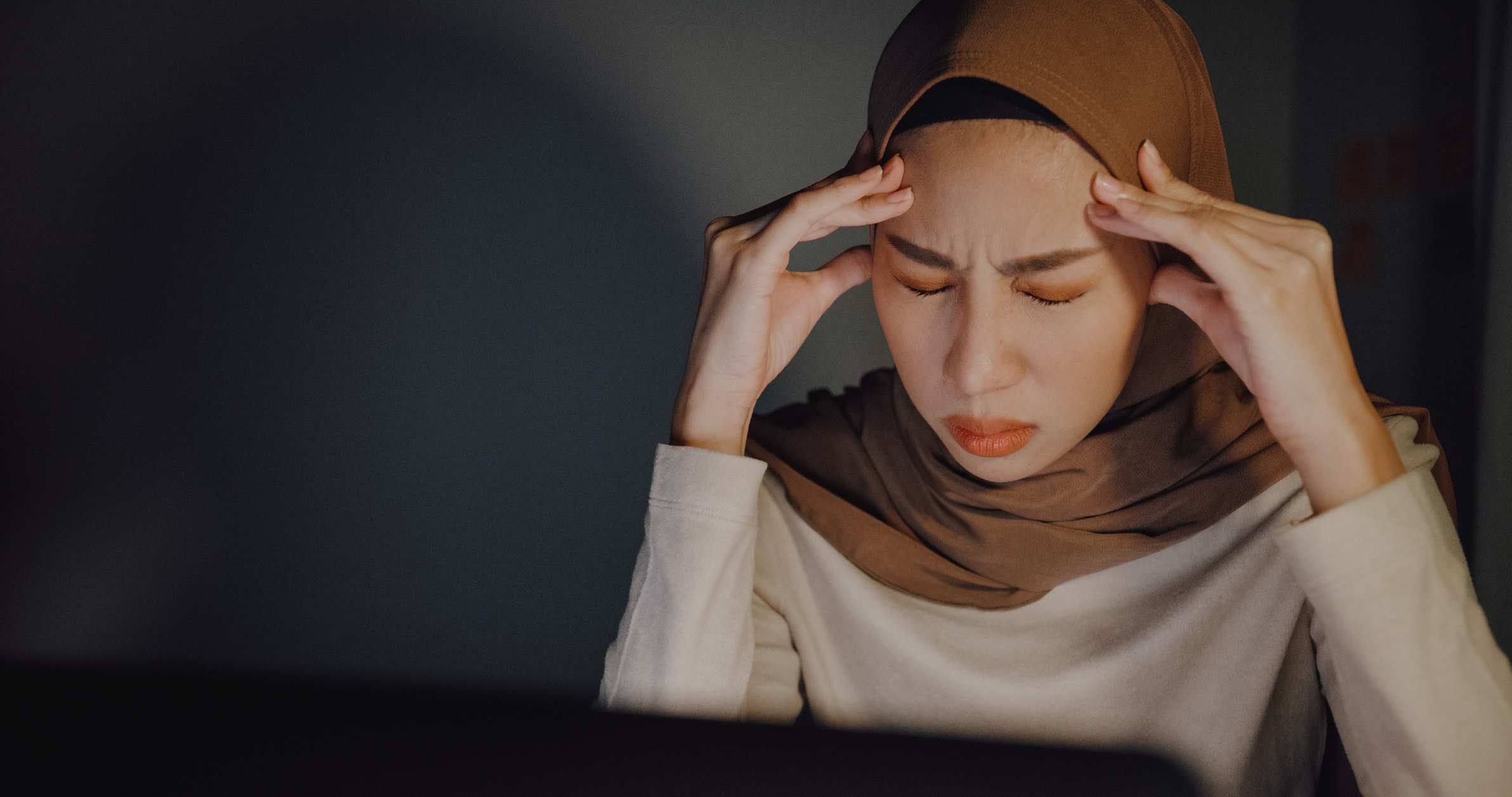 Woman in hijab appears stressed with hands on temples in front of a computer screen