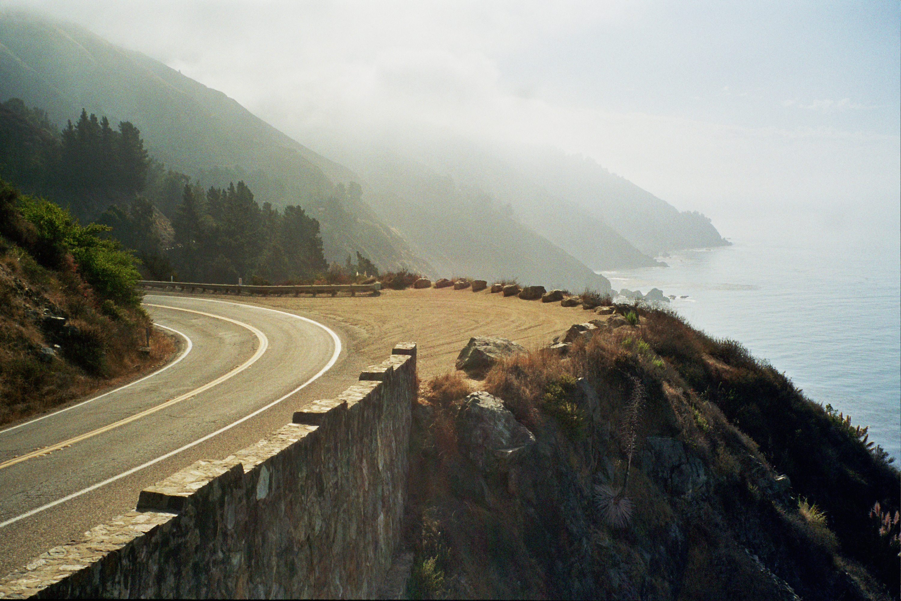 Curvy coastal road with a misty mountain backdrop and a calm ocean to one side