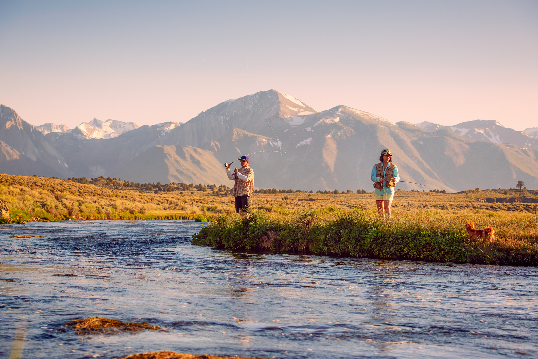 Two people fishing by a river with a mountain backdrop, one person gesturing