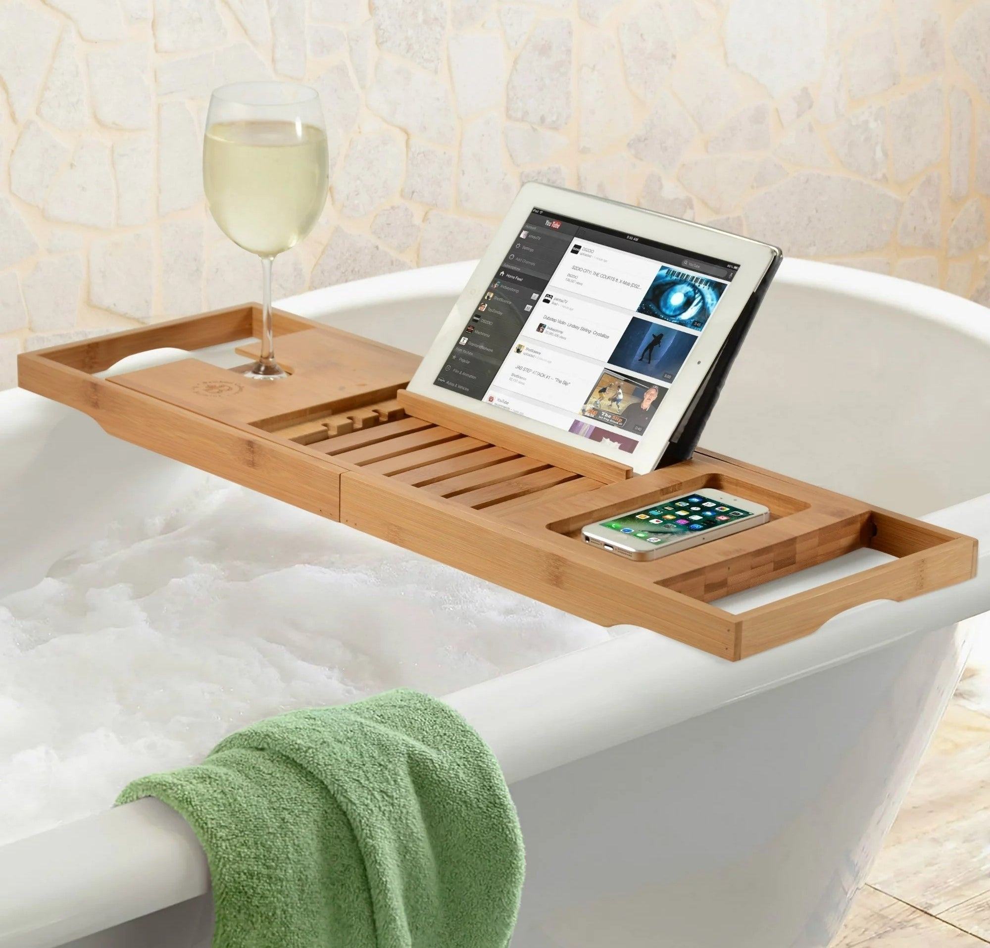 A wooden bath caddy across a bathtub holding a tablet, a phone, and a glass of wine