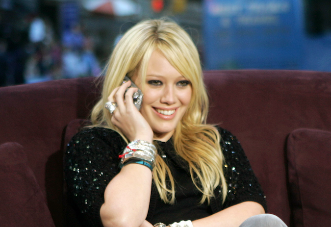 Hilary Duff smiling, seated, wearing a black sequined top, with numerous bracelets and holding a phone