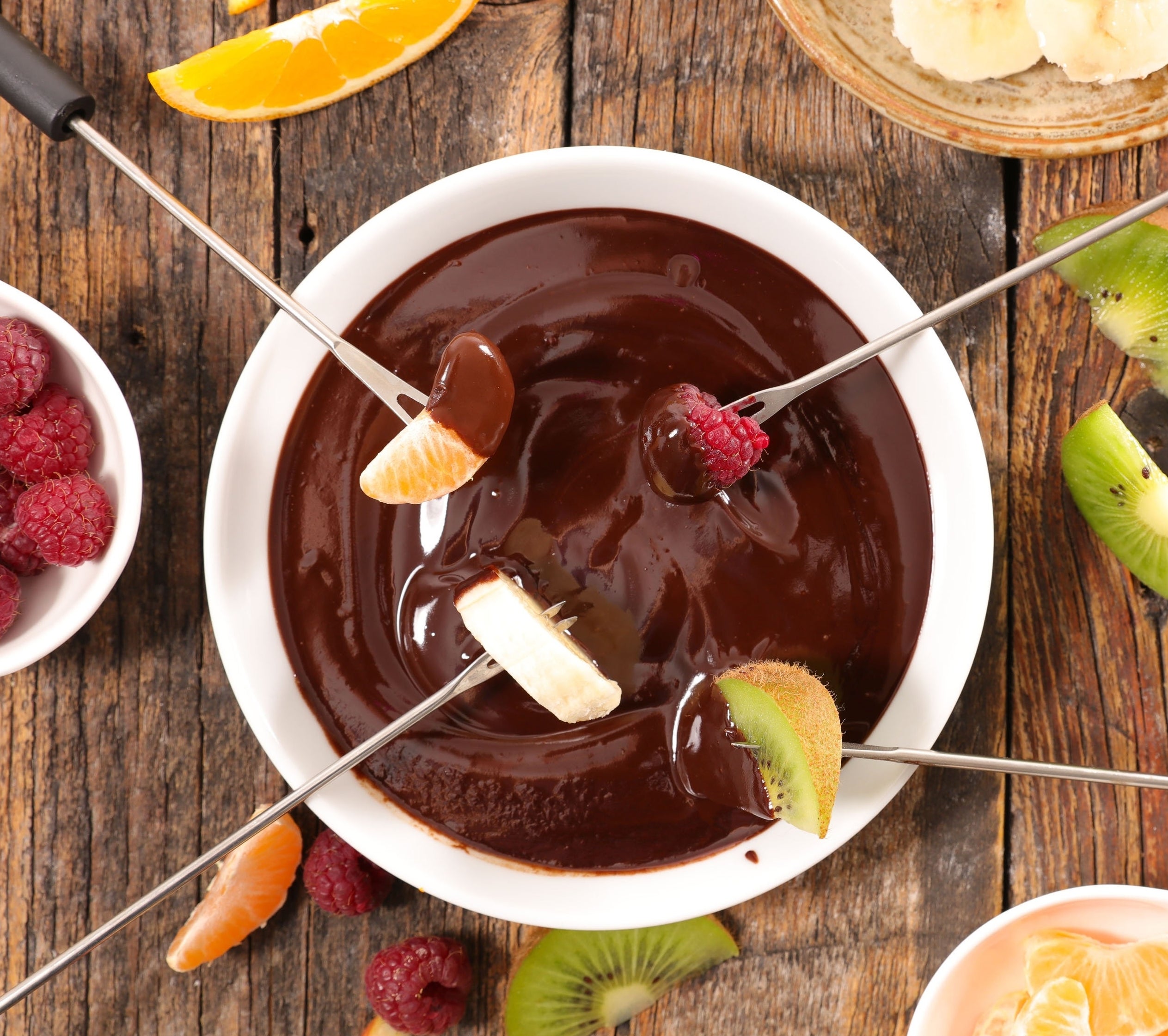 A bowl of chocolate fondue with fruits like banana, kiwi, and raspberry on skewers for dipping