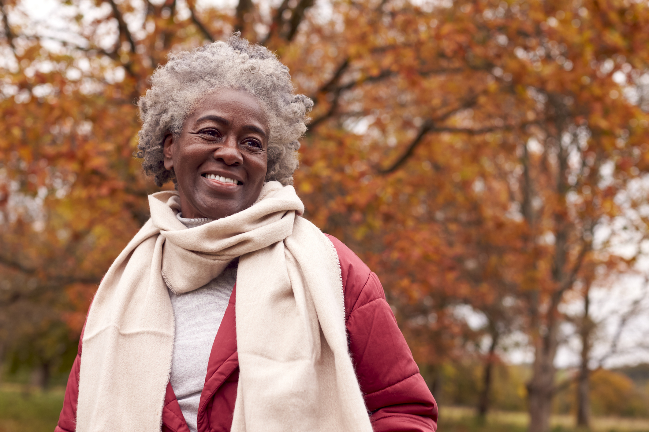 Smiling elderly woman with a scarf, outdoors with autumn trees in the background