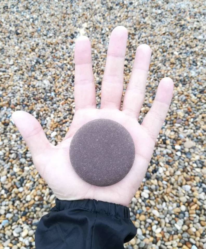 Open hand holding a round pebble, with a pebbly beach background