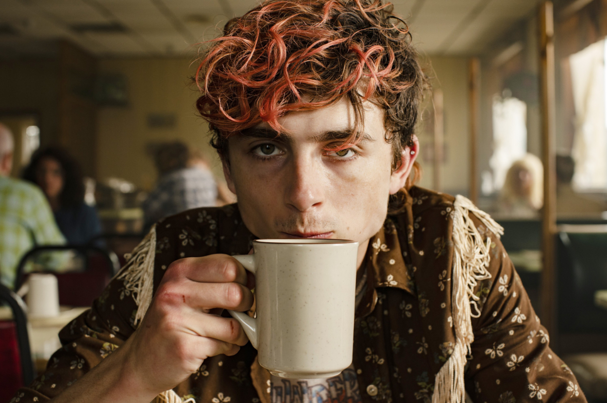 Timothee with orange-tinted hair sipping coffee, wearing a fringed jacket, in a diner. Other patrons in the background