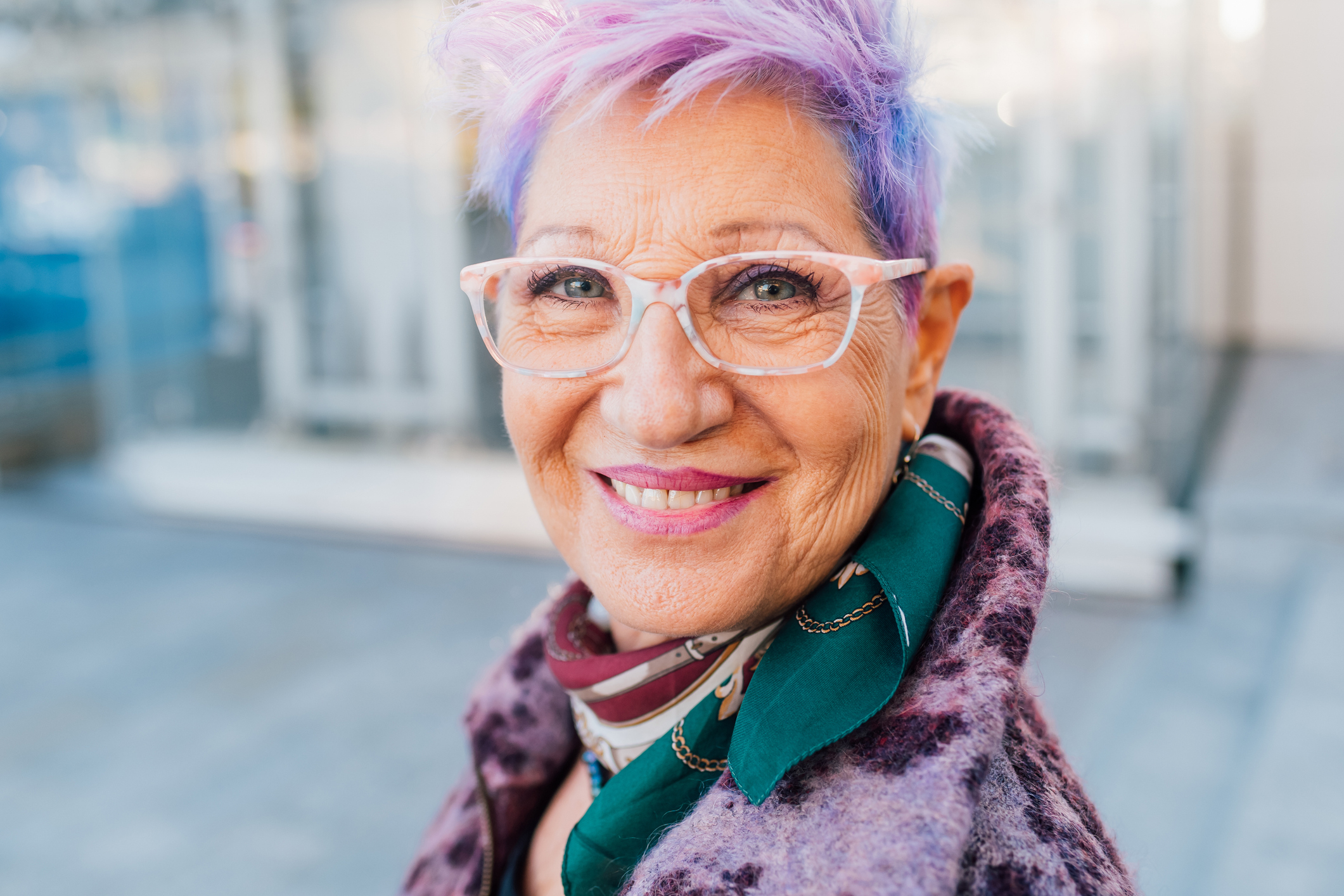 Elderly woman with purple hair wearing glasses and a scarf smiling at the camera