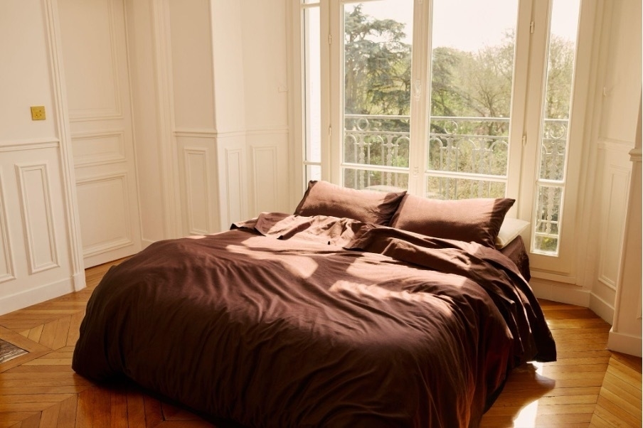 Unmade bed with dark bedding in a bright room with large windows and a view of greenery outside