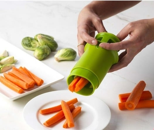 Hand using a green cylindrical kitchen gadget to chop carrots onto a plate