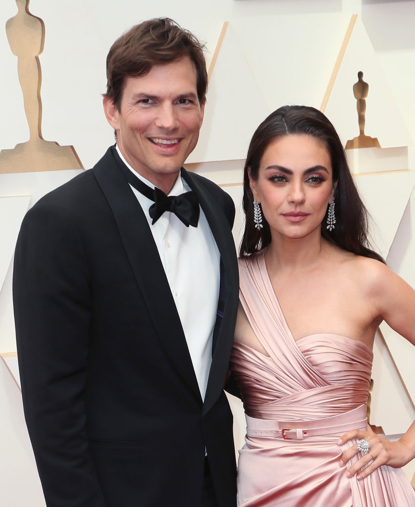 Two individuals posing together; one in a black tuxedo and the other in a draped pink gown with statement earrings