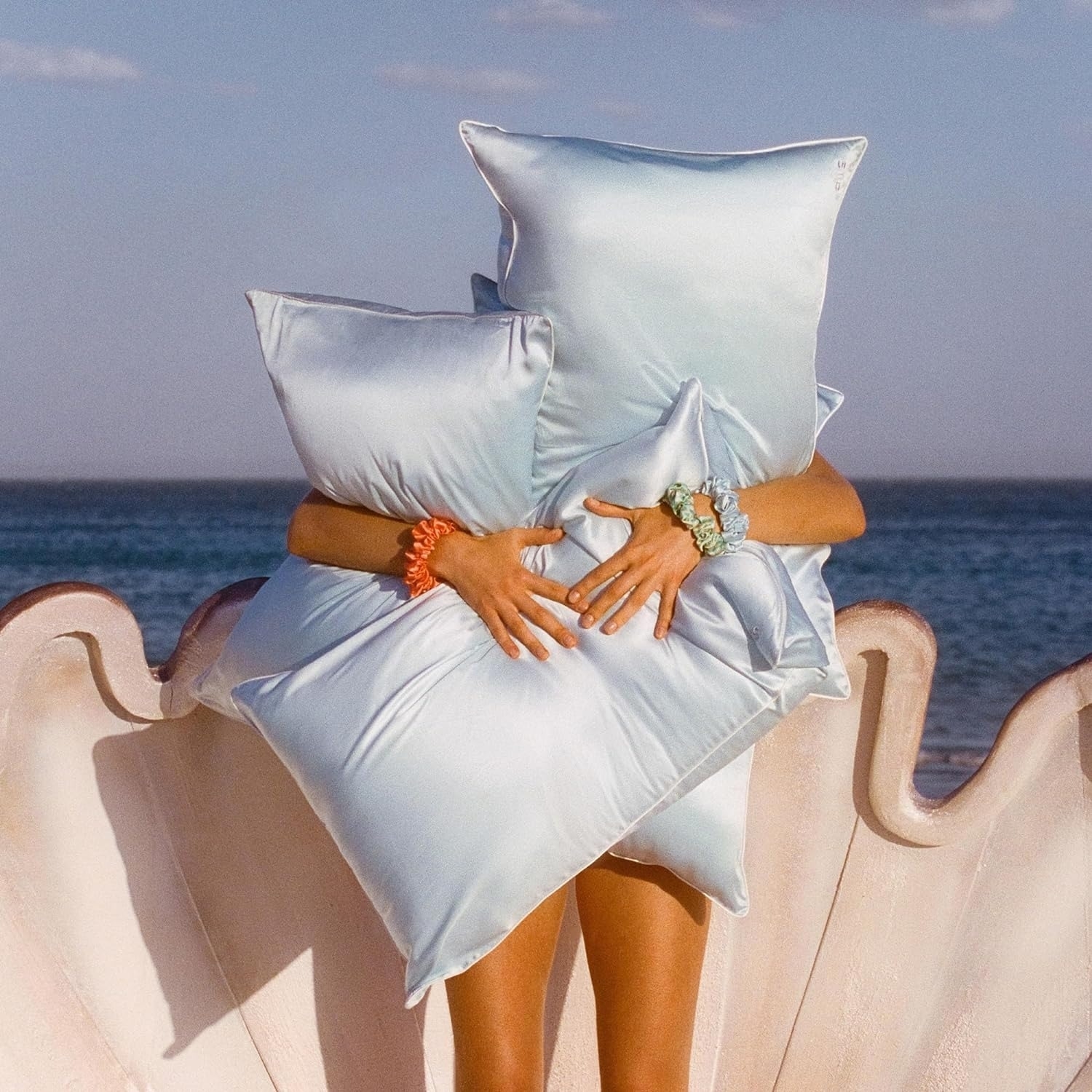Person holding large silk light blue pillows in front of them, with a seaside background