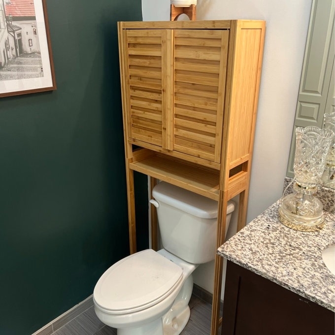 reviewer photo of the bamboo shelf over a white toilet, next to a sink with glassware on a marble countertop