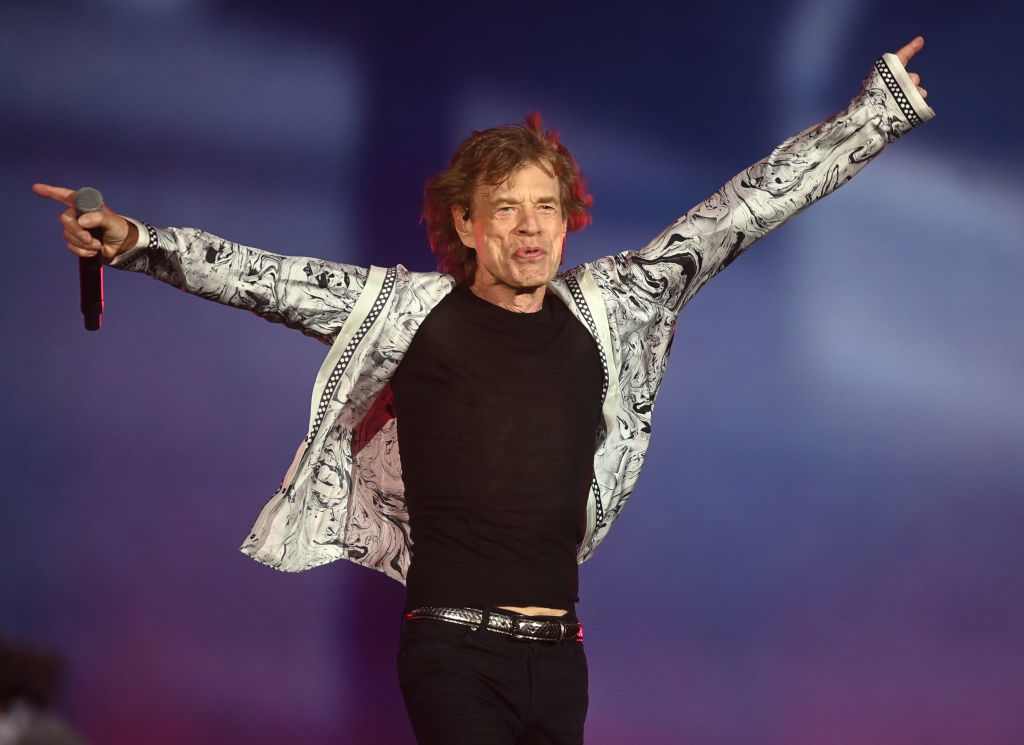 Person on stage with arms outstretched, wearing a patterned jacket and jeans