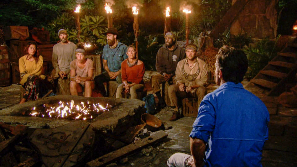 Group of contestants at night sitting around a fire during a Survivor Tribal Council