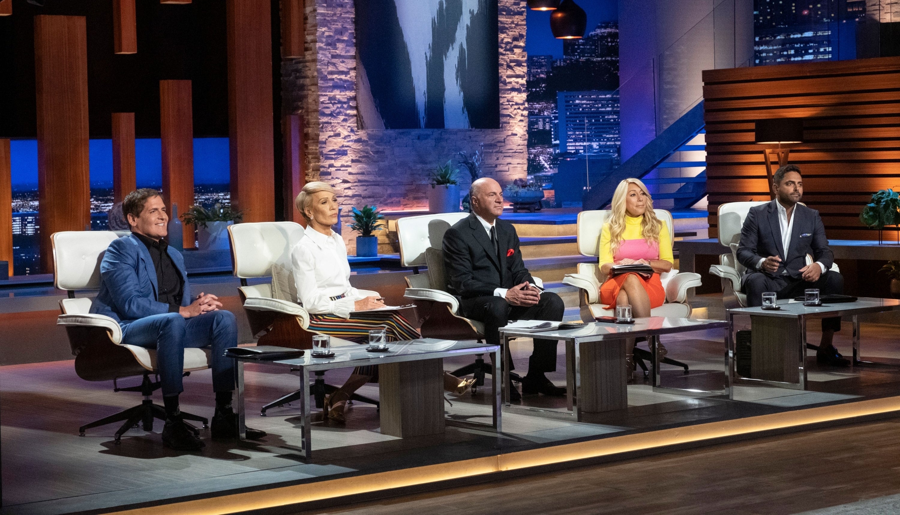 Five people seated in a row, on a TV show set designed to resemble a boardroom