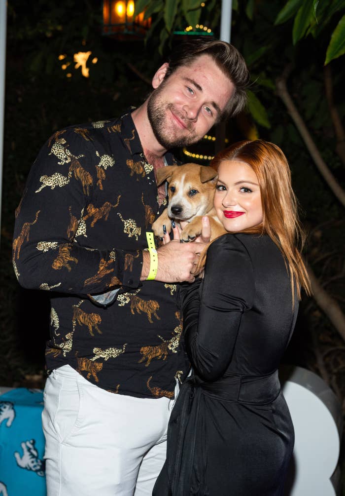 Two people smiling and embracing a small dog, man in patterned shirt, woman in black outfit