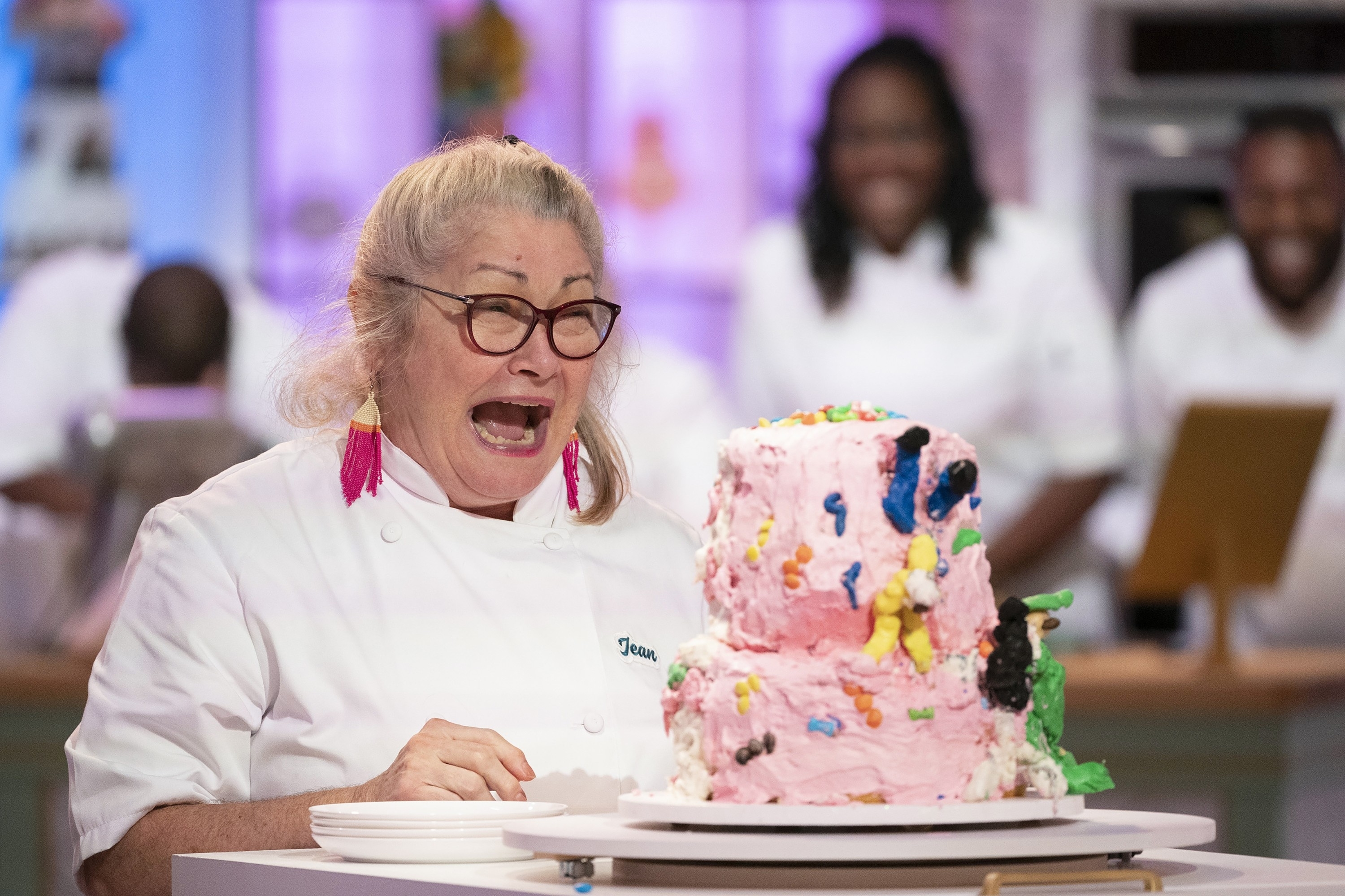 Woman in chef attire is surprised by a multi-tiered, colorful cake in front of her